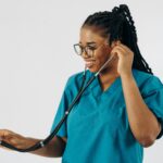 A black female doctor in blue scrubs, smiling while holding a stethoscope on a white background.