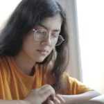 Low angle of diligent ethnic female student wearing casual t shirt and eyeglasses sitting at table with stacks of books and preparing for exam
