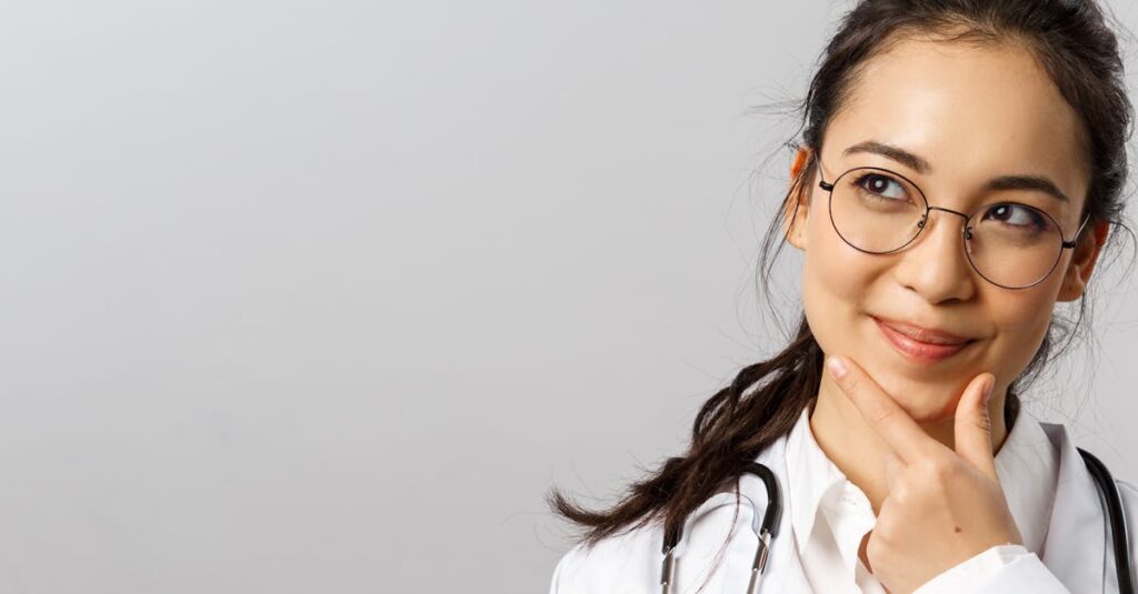 Asian female doctor wearing glasses, smiling thoughtfully with hand gesture on chin.