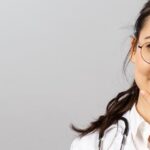 Asian female doctor wearing glasses, smiling thoughtfully with hand gesture on chin.