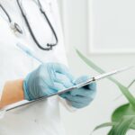 A healthcare professional wearing gloves takes notes on a clipboard with a stethoscope around their neck.