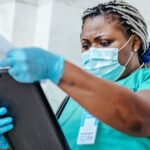Low angle of focused black female nurse in mask and gloves standing in hospital and looking through papers