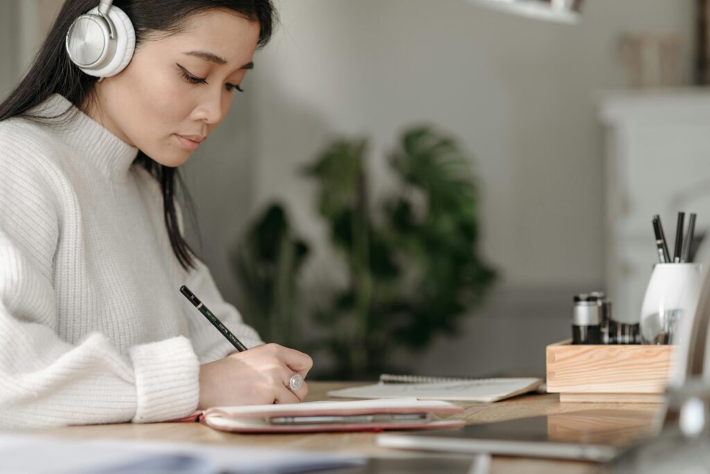 Asian woman wearing headphones writing at home, concentrating on her notes.