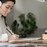Asian woman wearing headphones writing at home, concentrating on her notes.