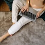 Pregnant woman sitting on carpet working remotely on laptop, enjoying a comfortable home environment.