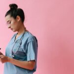 Female nurse in scrubs writing on a clipboard with a stethoscope on a pink background.