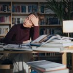 A tired woman surrounded by books, studying in a library, feeling overworked and stressed.