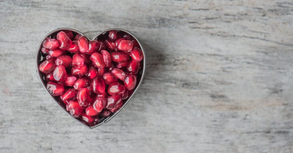 Delicious and juicy pomegranate seeds in a heart-shaped bowl on a wooden table.